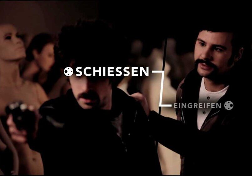 Zwei Männer sind von Schaufensterpuppen umgeben; einer hält eine Waffe, während der andere seine Schulter berührt. Der überlagerte Text auf Deutsch lautet "SCHIESSEN" und "EINGREIFEN", jeweils mit einem Zielsymbol.