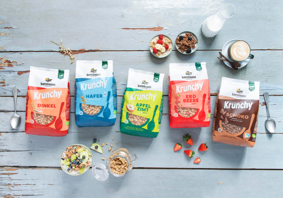 Fünf Tüten Krunchy-Müsli in verschiedenen Geschmacksrichtungen stehen in einer Reihe auf einem hellblauen Holztisch, umgeben von Schalen mit Müsli und Früchten, einem Cappuccino, Milch und zwei Löffeln.