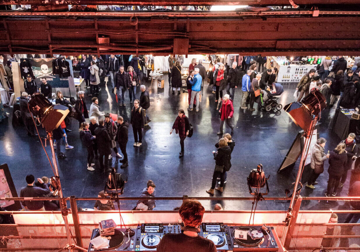 Ein DJ steht an einem Mischpult mit Plattenspielern und Geräten und tritt auf einer erhöhten Plattform auf, von der aus er eine große Menschenmenge überblickt, die sich bei einer Veranstaltung oder Ausstellung in einem Innenraum versammelt hat.