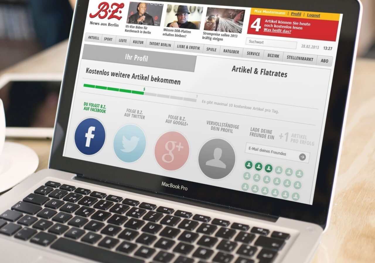 Ein MacBook Pro zeigt eine deutschsprachige Nachrichten-Website mit Social-Media-Symbolen für Facebook, Twitter, Google+ und E-Mail an und lädt Nutzer ein, ihr zu folgen oder sie zu teilen, um Zugang zu weiteren Artikeln zu erhalten. Auf der rechten Seite ist teilweise eine Kaffeetasse zu sehen.