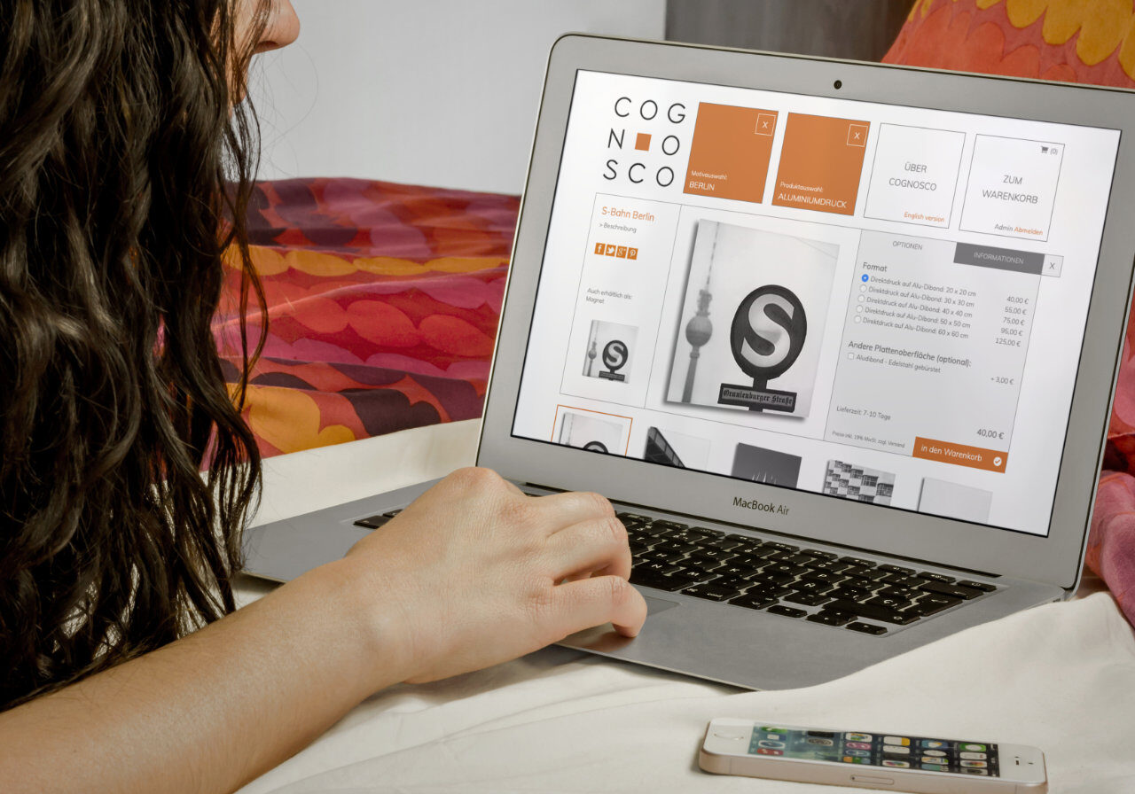 Eine Person sitzt mit einem MacBook Air Laptop auf einem Bett und stöbert in einem Online-Shop namens Cognosco; ein Smartphone liegt in der Nähe auf dem Bett. Auf dem Bildschirm werden Produkte und Preise angezeigt.