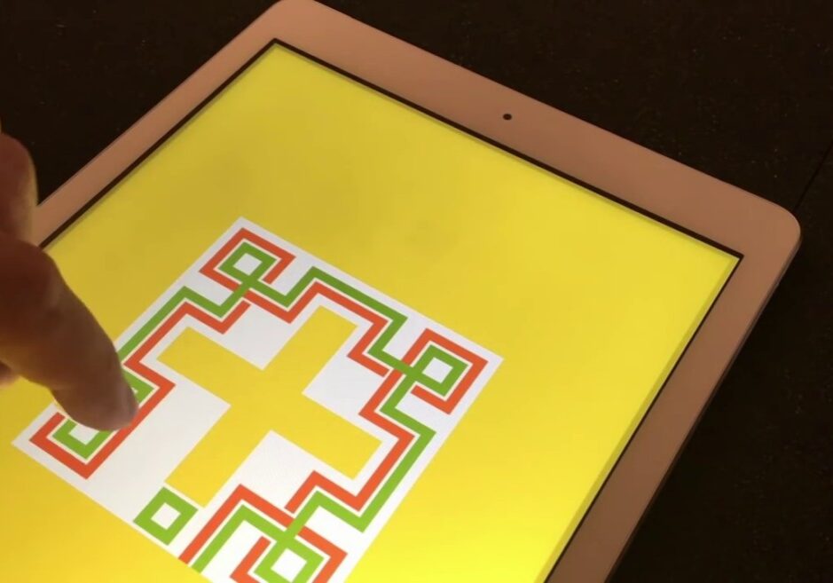 Eine Hand zeigt auf einen Tablet-Bildschirm, der einen gelben Hintergrund mit einem geometrischen Muster zeigt, das ein gelbes Kreuz mit grünen, roten und weißen Umrissen darstellt.