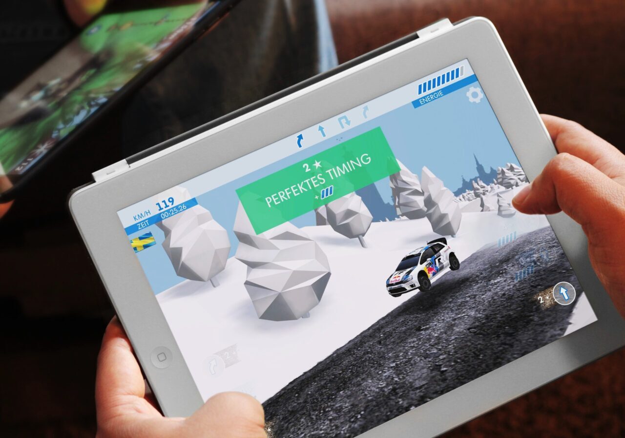 Eine Person hält ein weißes Tablet und spielt ein Rennspiel mit einer Schneelandschaft und einem Rallyeauto auf dem Bildschirm. Die Phrase Perfektes Timing wird auf der Spieloberfläche angezeigt.