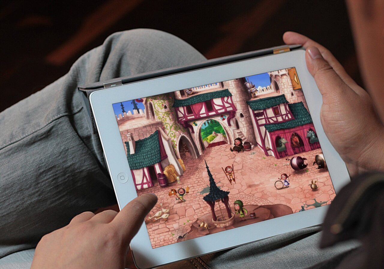 Eine Person hält ein Tablet auf dem Schoß und spielt ein farbenfrohes mittelalterliches Videospiel mit einer Burg, Kopfsteinpflasterwegen und animierten Figuren. Die Person interagiert mit einem Finger auf dem Touchscreen.